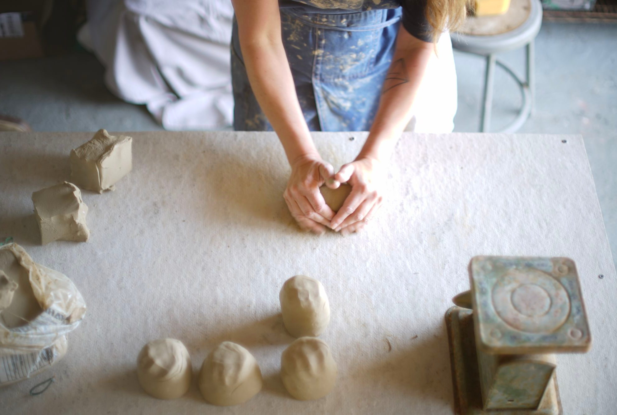 Top-down view of a person in a black shirt and blue apron working with clay on a white work surface, with clay sculptures and tools around.