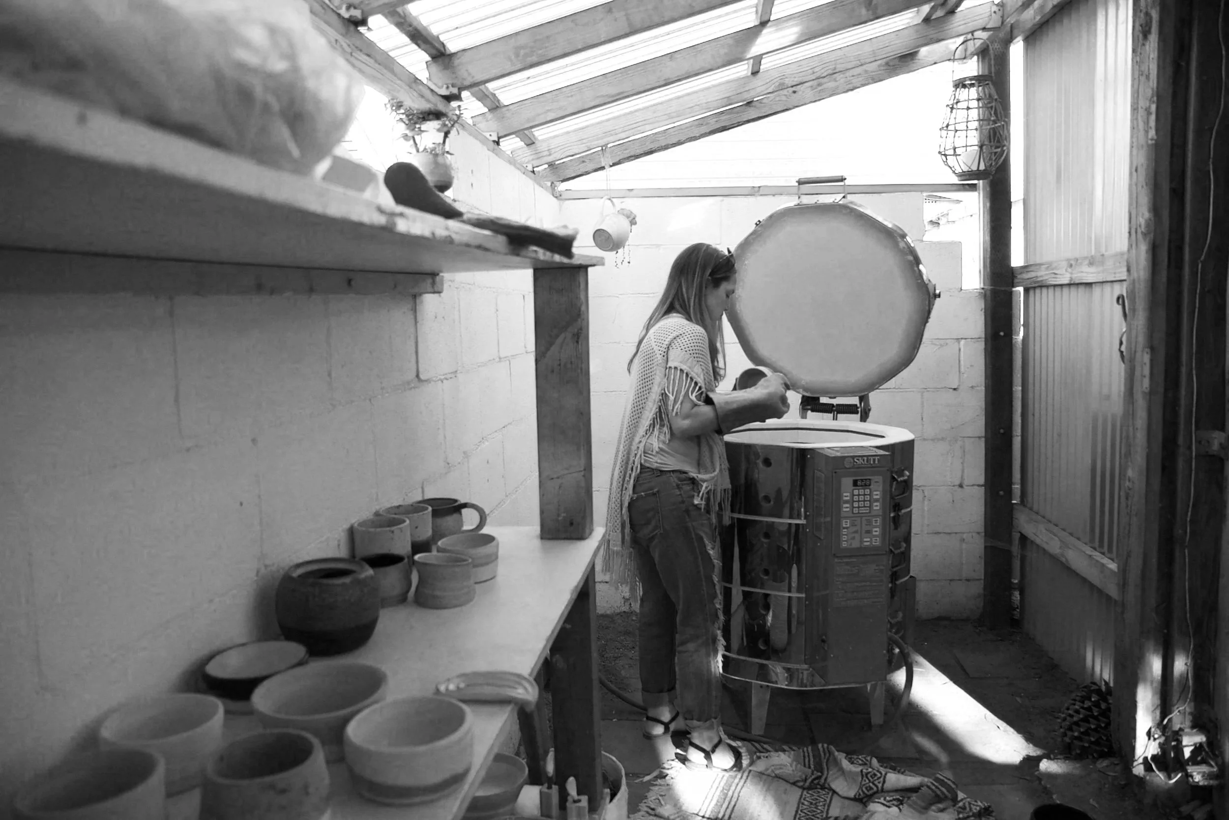 A woman standing in a rustic workshop or pottery studio, working with clay on a pottery wheel. The workspace has wooden shelves with bowls and pottery items, and the room is constructed with wooden beams and corrugated metal walls. Sunlight streams in through a window.