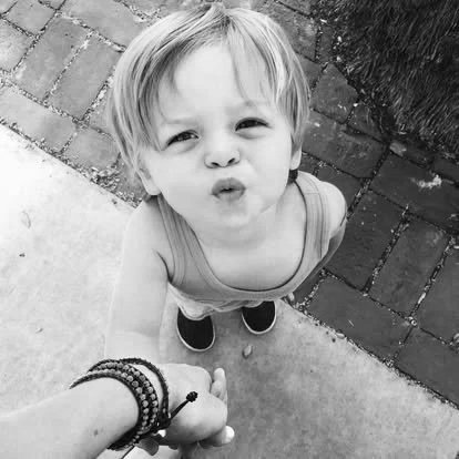 A young boy with blonde hair making a pouty face, standing outdoors on a sidewalk, holding an adult's hand.