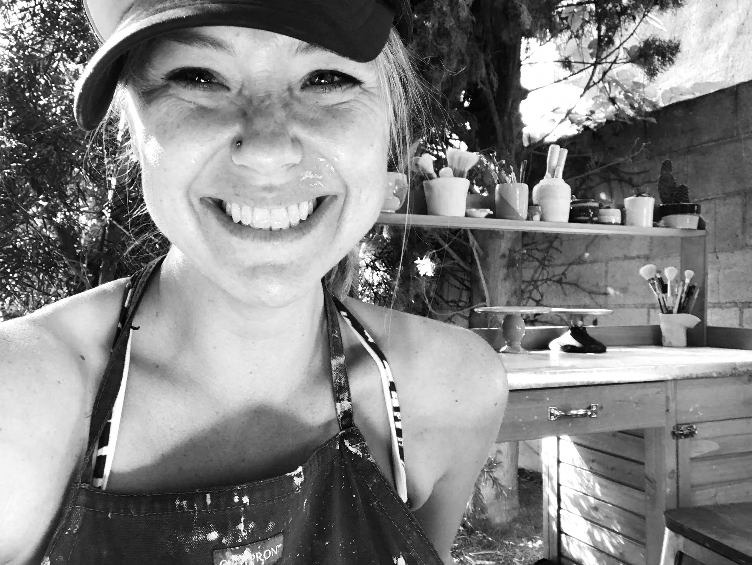 A woman smiling outdoors, wearing a baseball cap and a patterned apron, with gardening tools and potted plants on shelves in the background.