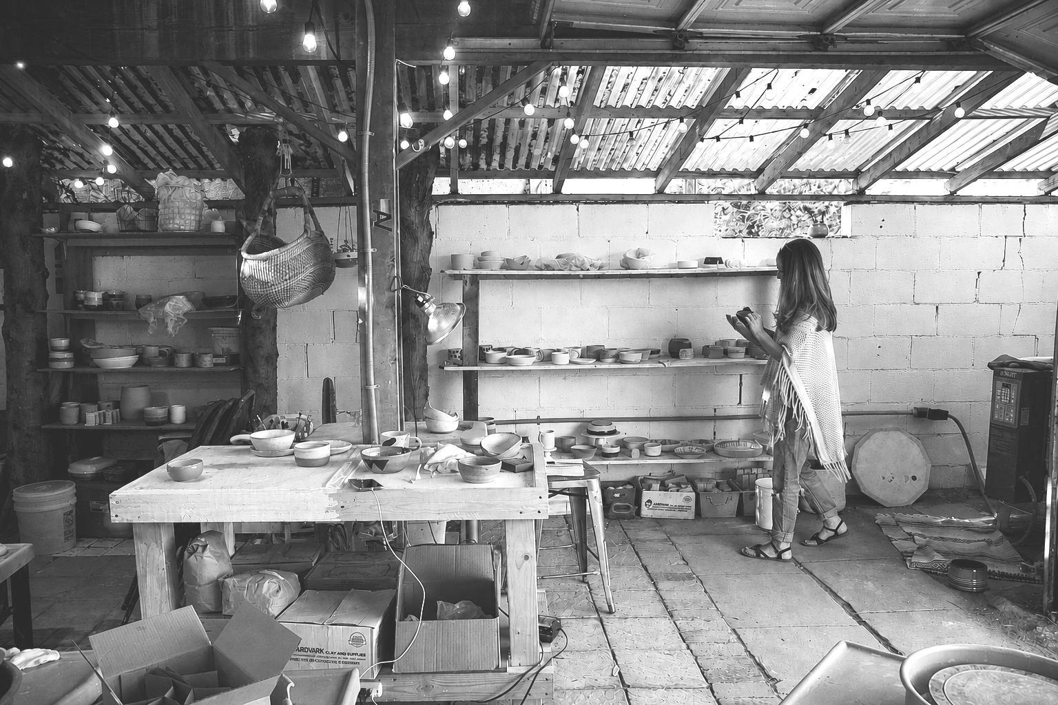 Black and white photo of a woman in a pottery workshop, surrounded by shelves filled with ceramic bowls and crafts, working on her pottery projects.