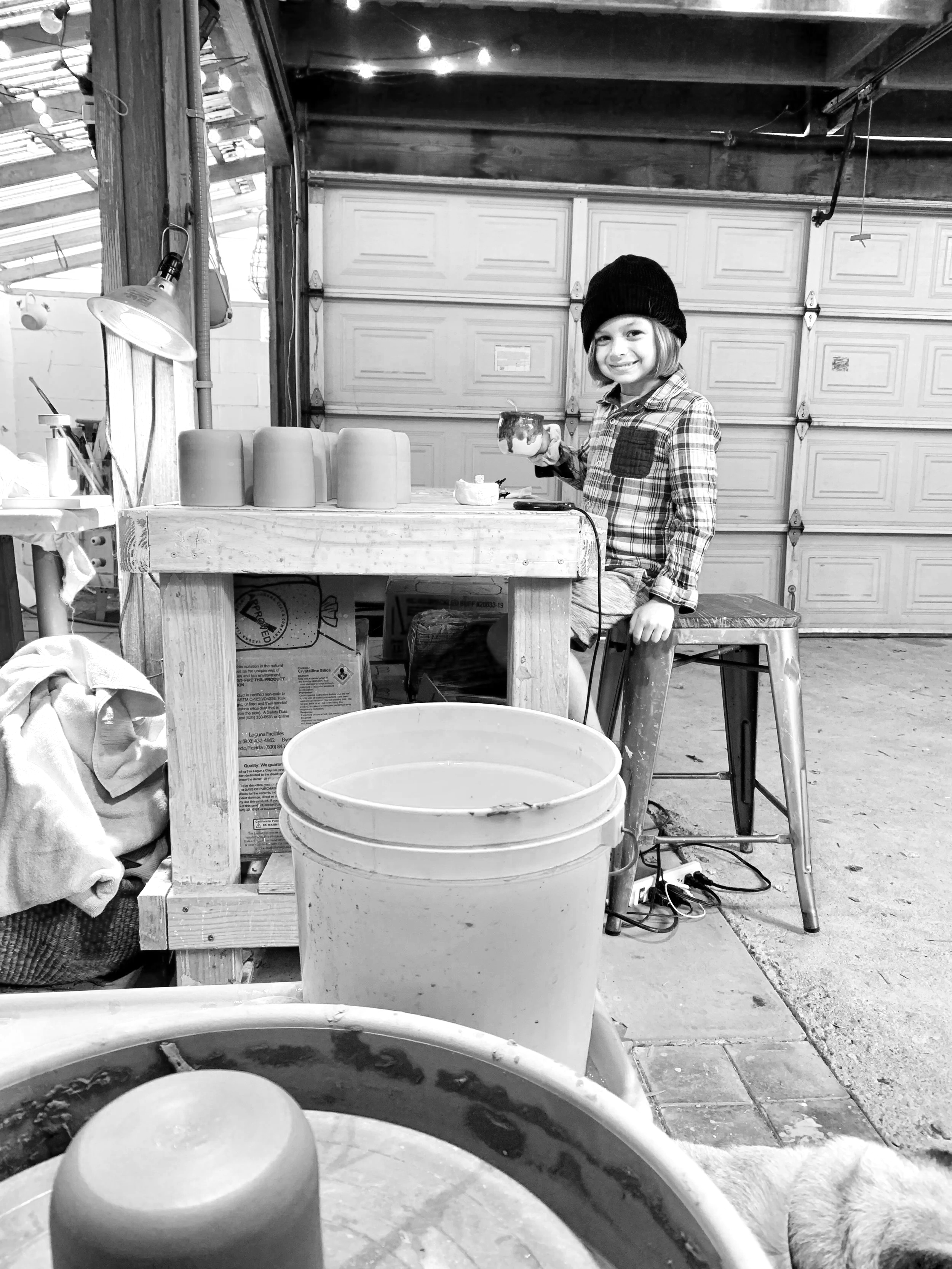 A young girl sits on a stool in a garage, smiling and holding a mug. The garage door is closed, and there are various tools and objects like cups, buckets, and wires around her. The scene is in black and white.
