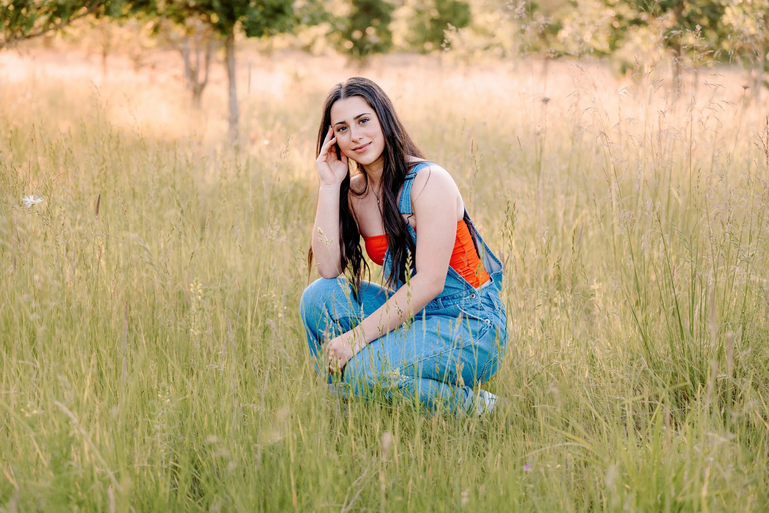 Girl squatting in field at sunset for senior photos wearing a red shirt and overalls