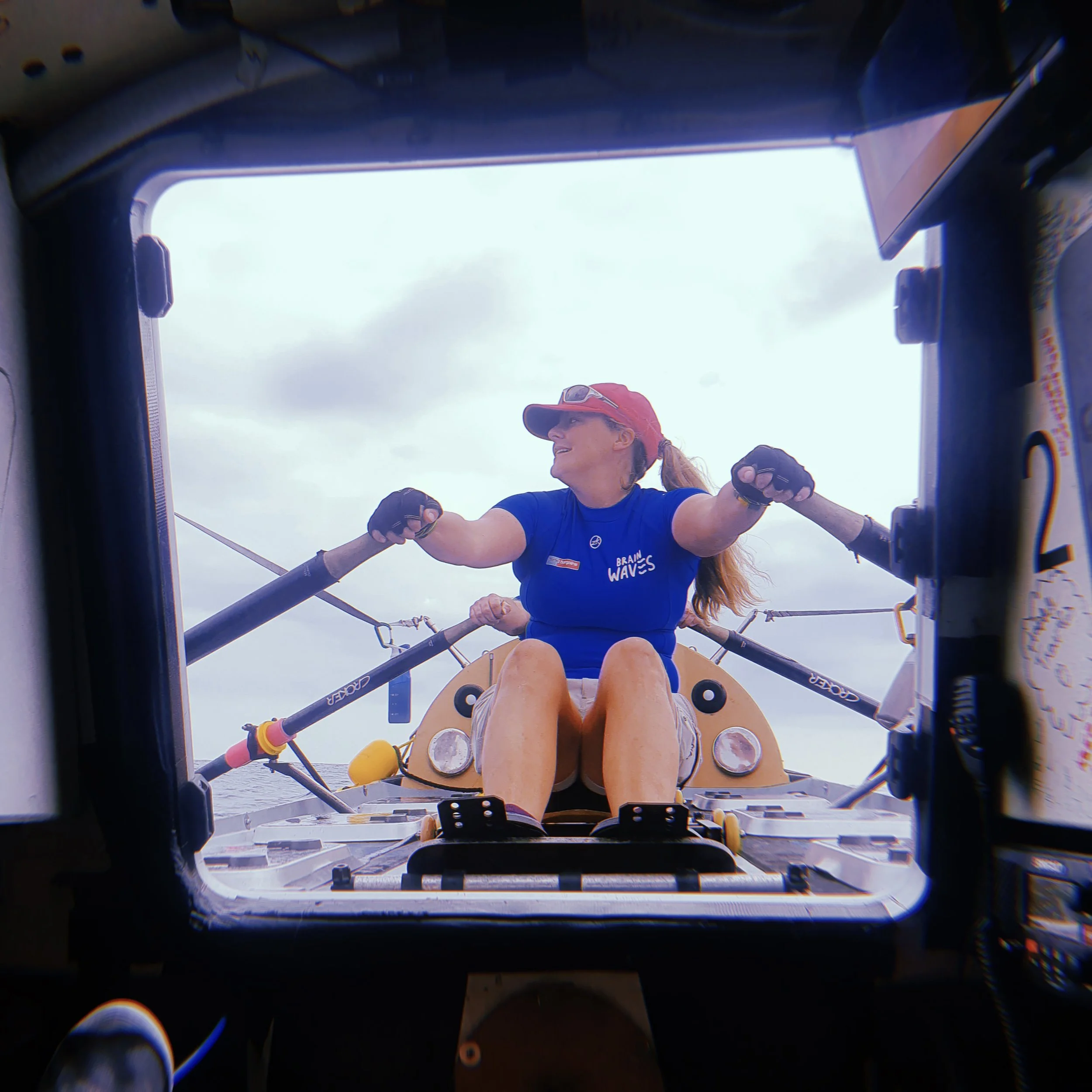 A woman in a blue shirt and red cap is rowing a boat from a view inside the boat's cockpit, with oars extended outward. She is smiling and looking to her left. The sky is cloudy.