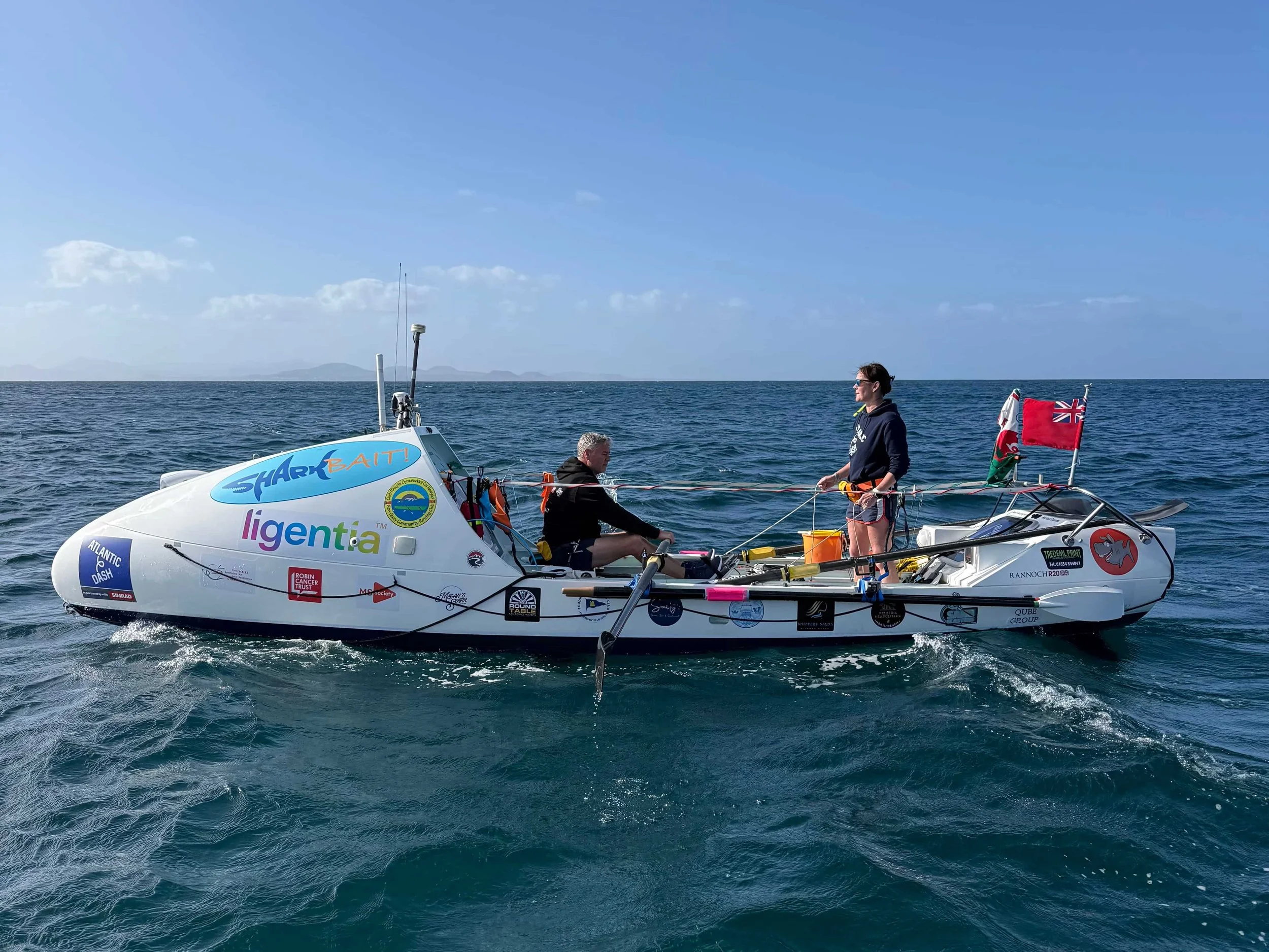 Two people in a small ocean rowing boat covered in sponsor logos, floating on the ocean under a clear blue sky.