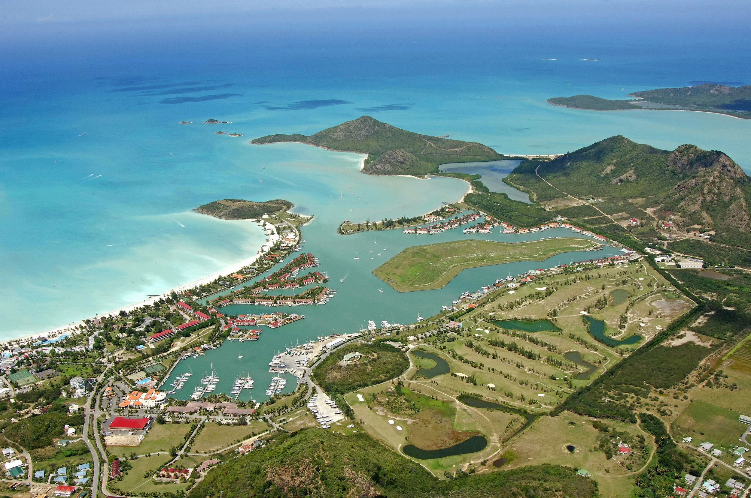 Aerial view of a luxury resort with marina, beaches, green landscapes, and surrounding hills and mountains, with turquoise ocean in the background.