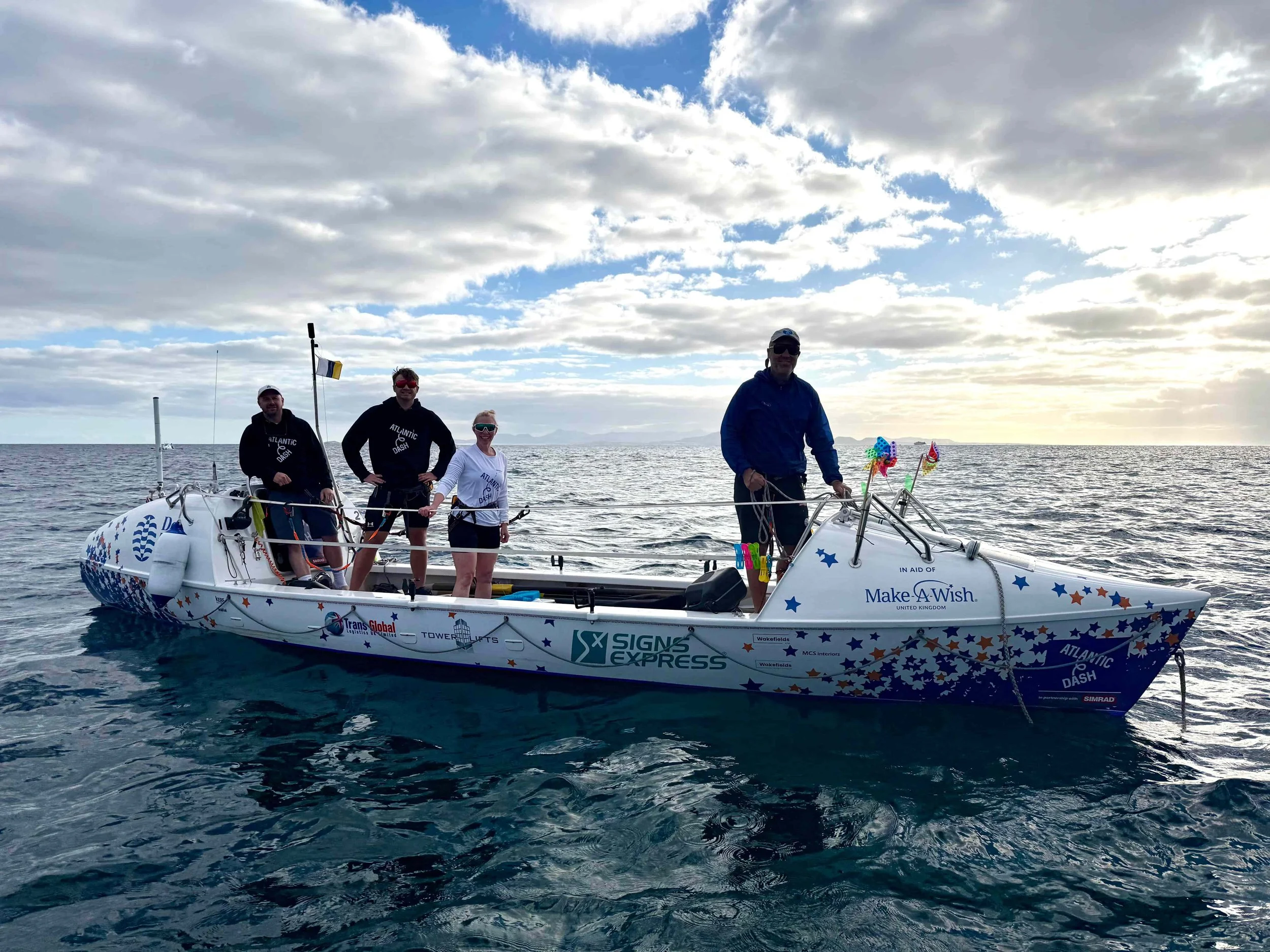 four people in a small ocean rowing boat covered in sponsor logos, floating on the ocean under a clear blue sky.