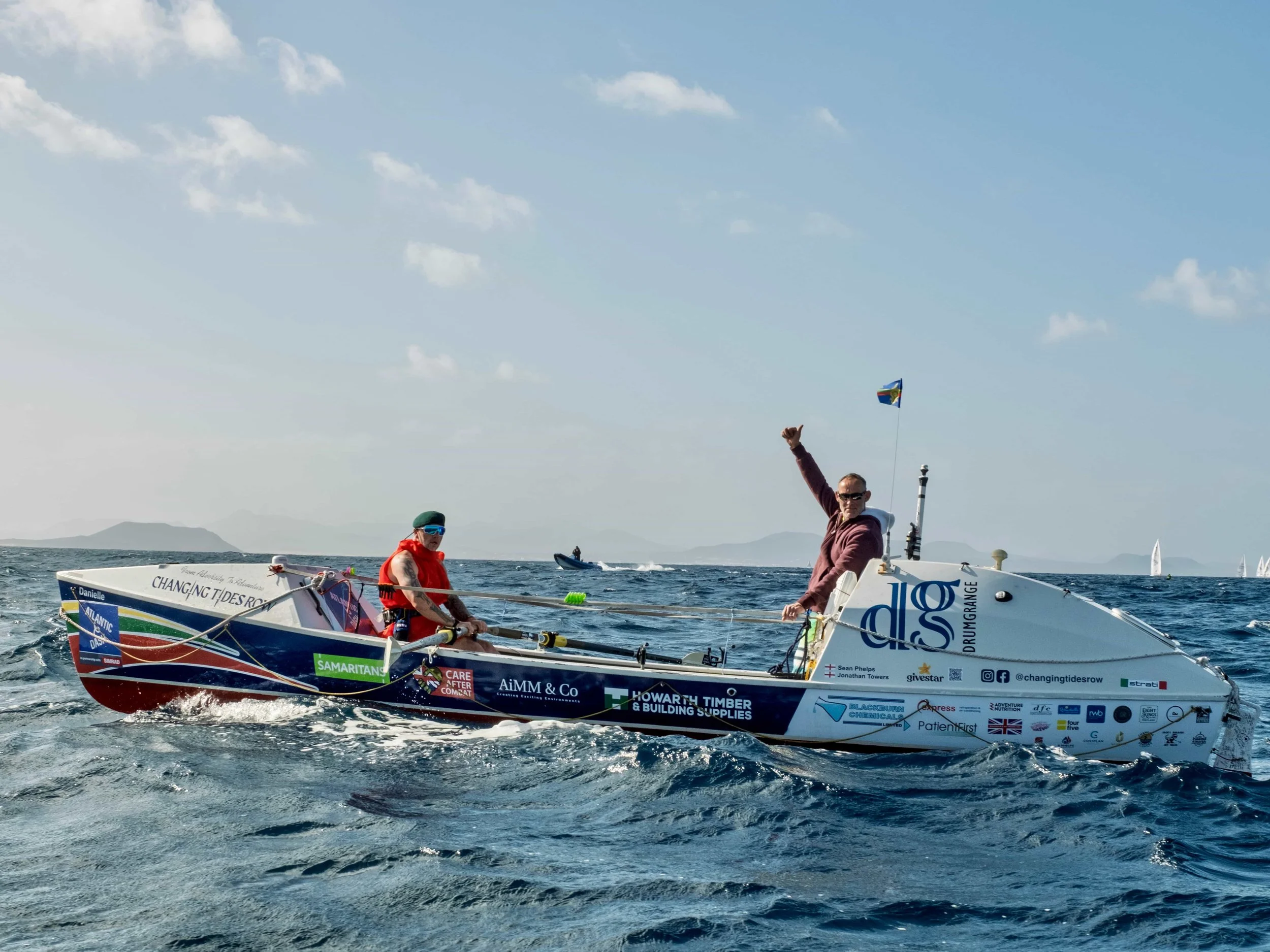 Two people on a small ocean rowing boat covered in logos, floating on the ocean under a clear blue sky.