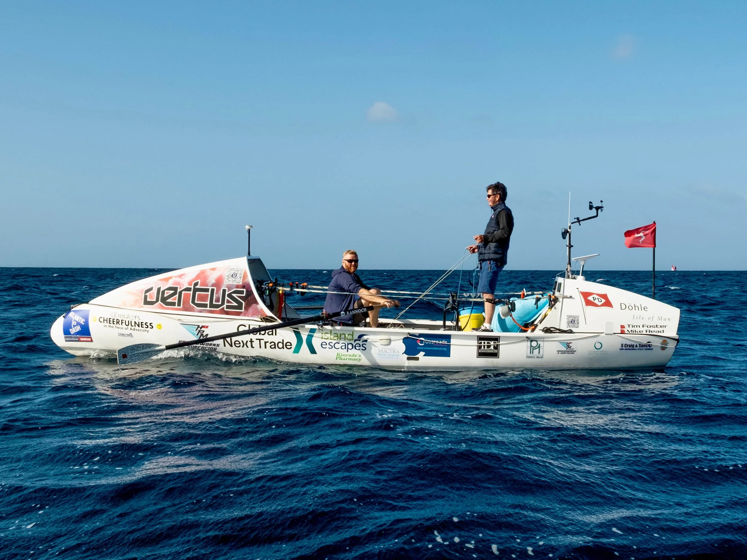 Two people in a small ocean rowing boat covered in sponsor logos, floating on the ocean under a clear blue sky.