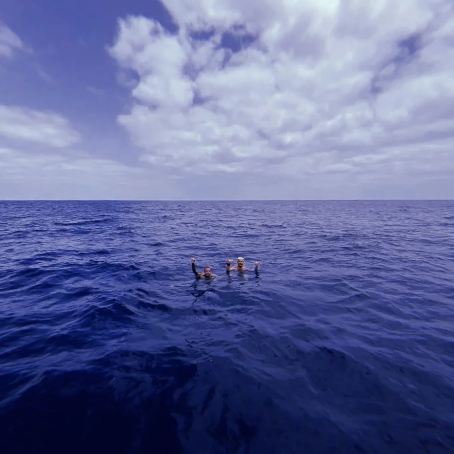 Two people swimming in the open ocean on a sunny day with partly cloudy sky.