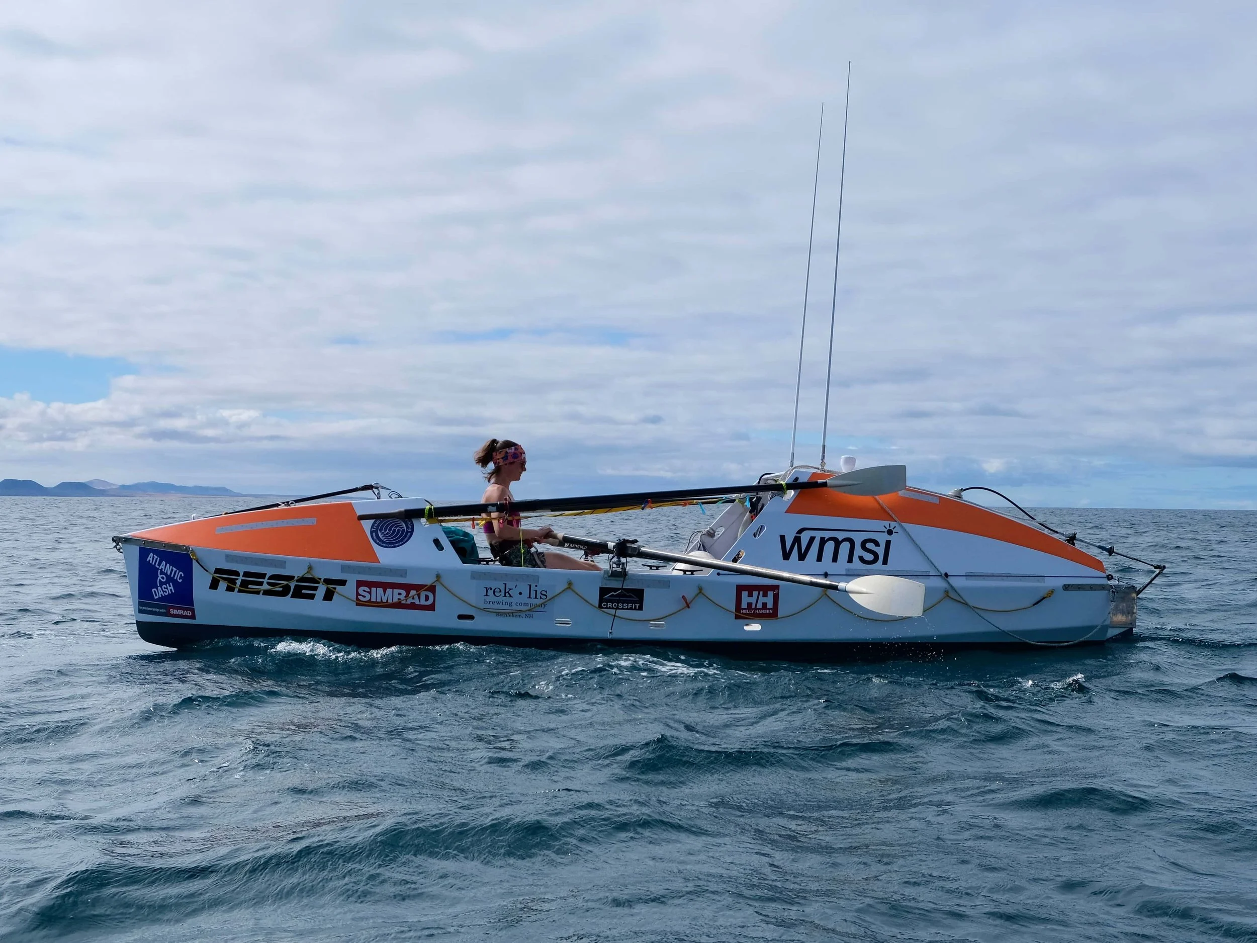 one female in a small ocean rowing boat covered in sponsor logos, floating on the ocean under a clear blue sky.