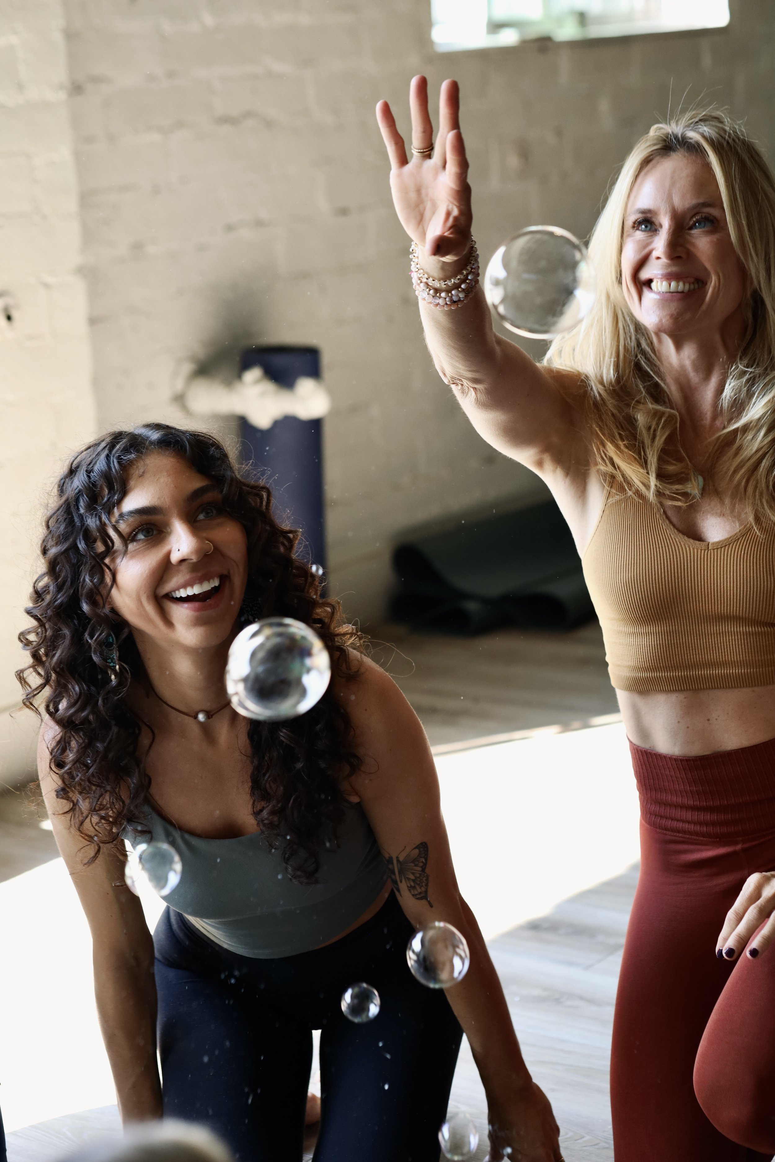 Two women playing with bubbles indoors, one kneeling and one standing, smiling and reaching to pop bubbles