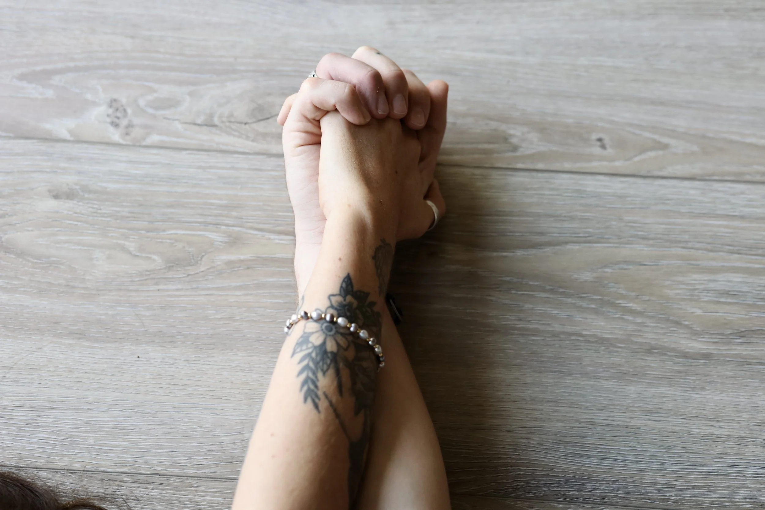 Hands clasped on a wooden surface, one forearm with a floral tattoo and a bracelet.