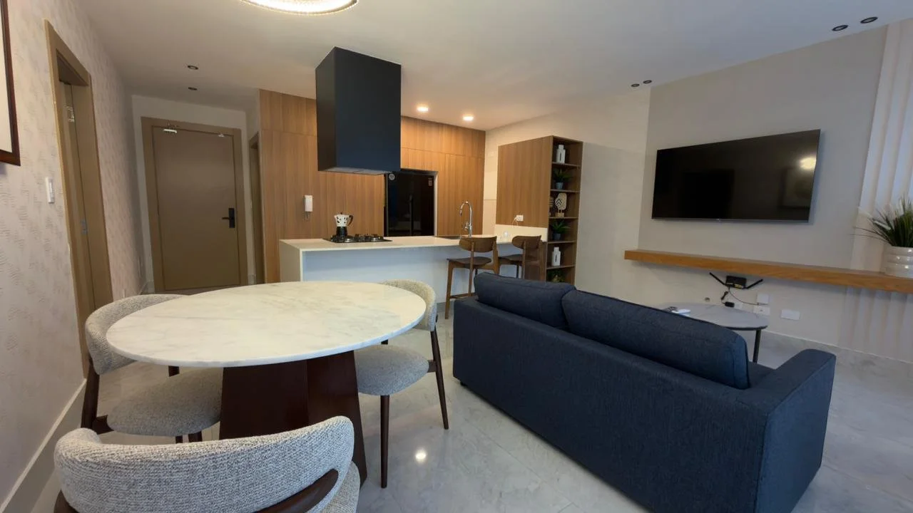 Modern living room with a sofa facing a wall-mounted TV, open kitchen with a breakfast bar, dining table with four chairs, and a bookshelf on the back wall.