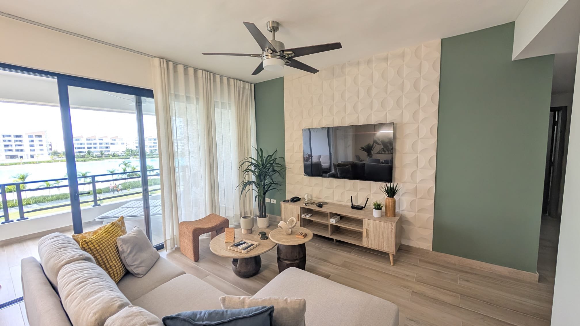 Living room with a sliding glass door showing an outdoor view, a beige sectional sofa with cushions, a wooden TV stand with a flat-screen TV, decorative plants, and a ceiling fan.