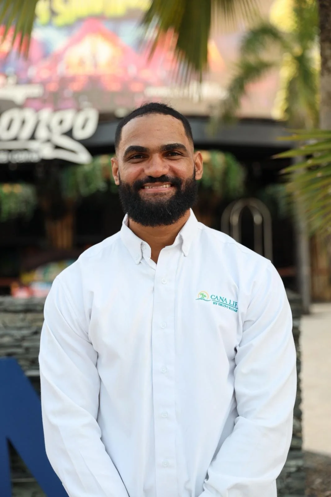 A man in a suit sitting on a wooden bench made from logs, under a thatched roof, with a railing and street scene in the background.