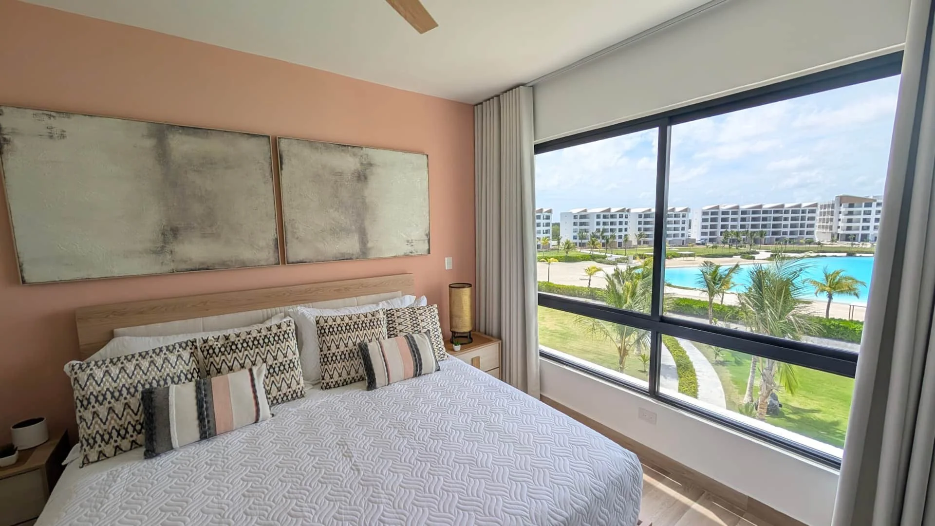 Bedroom with a large bed, decorative pillows, a building with a pool and palm trees outside the window.