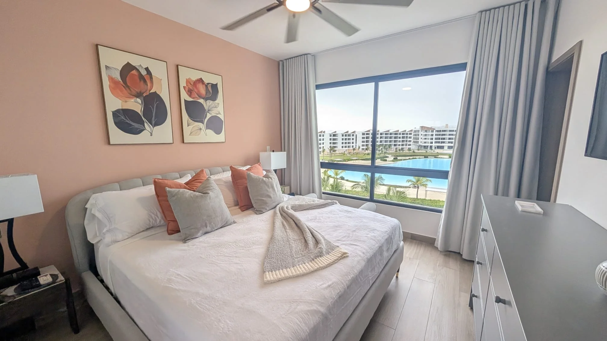 Bedroom with a large bed, pillows, and a blanket, two side tables with lamps, large window showing a view of a pool and modern buildings outside.