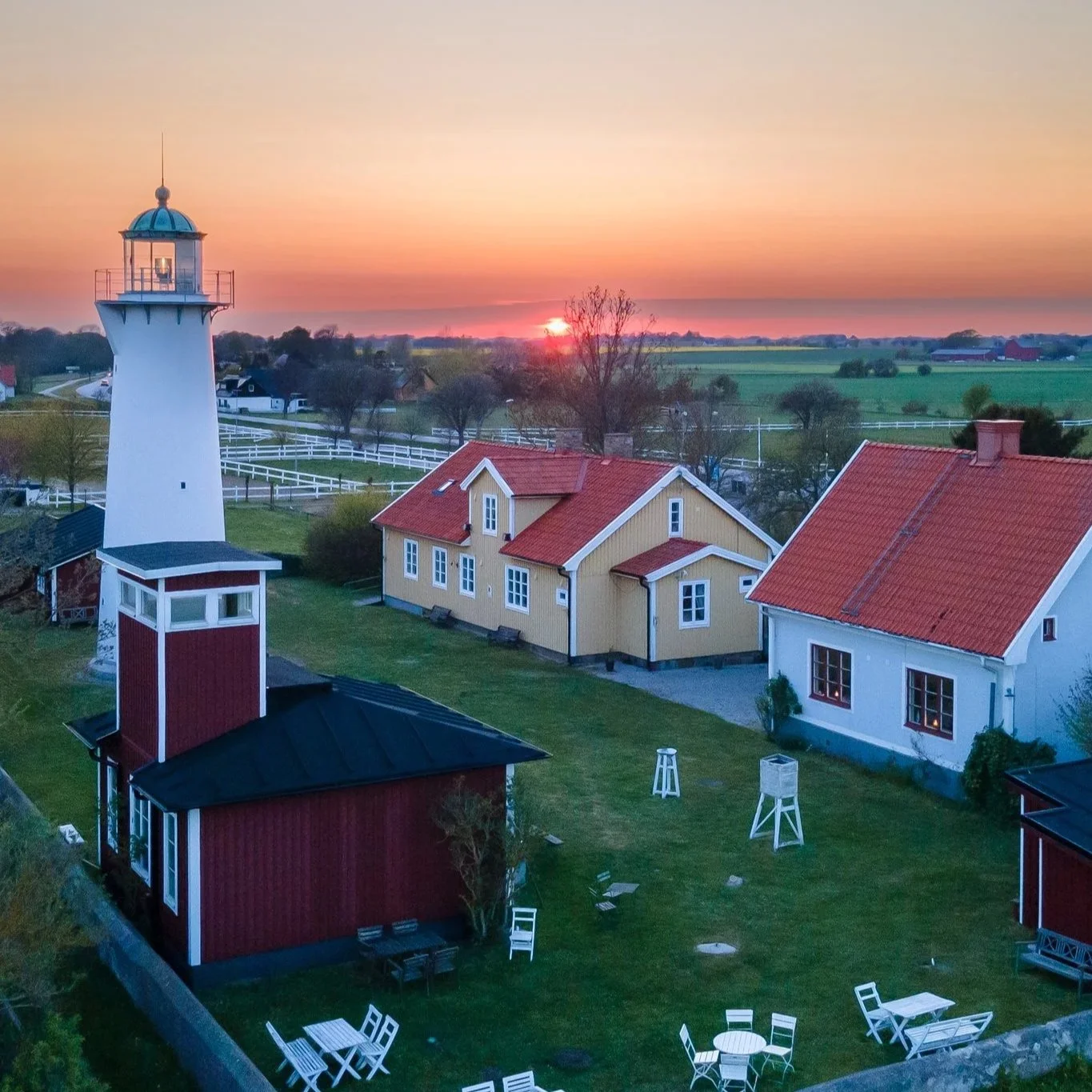 A scenic view of a lighthouse and surrounding houses during sunset, with green fields in the background.