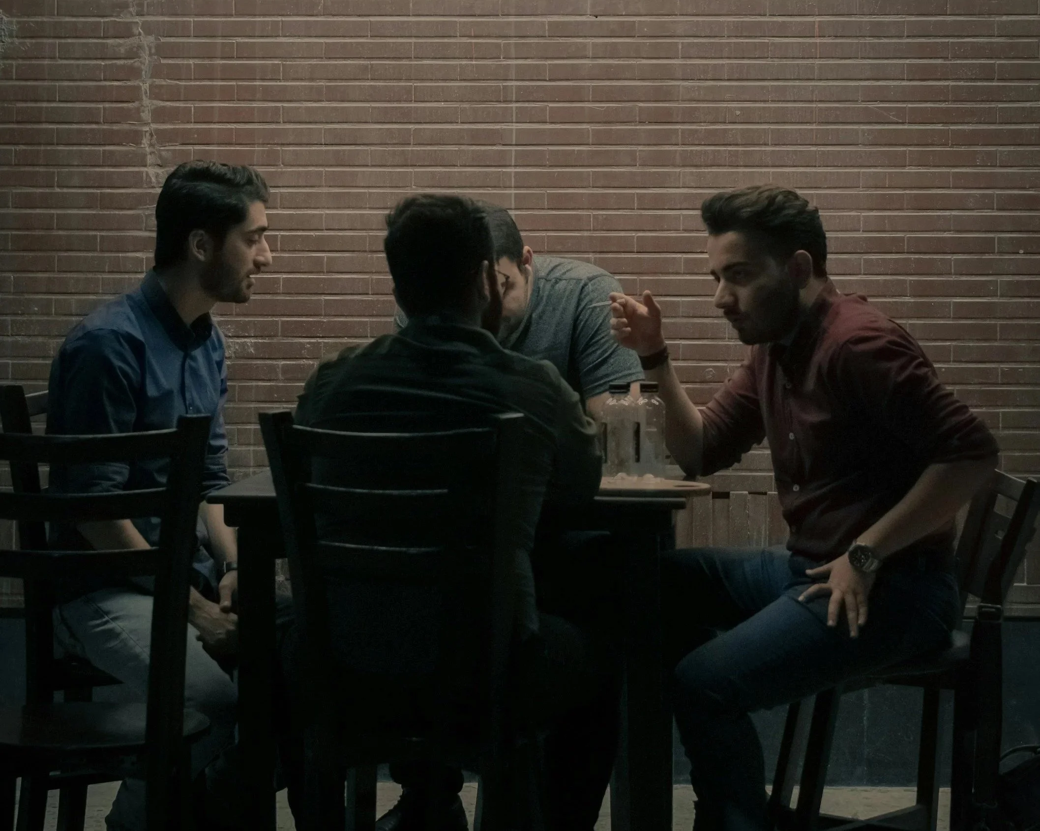 Five young men sitting around a table in a dimly lit room with a brick wall background, engaging in conversation.