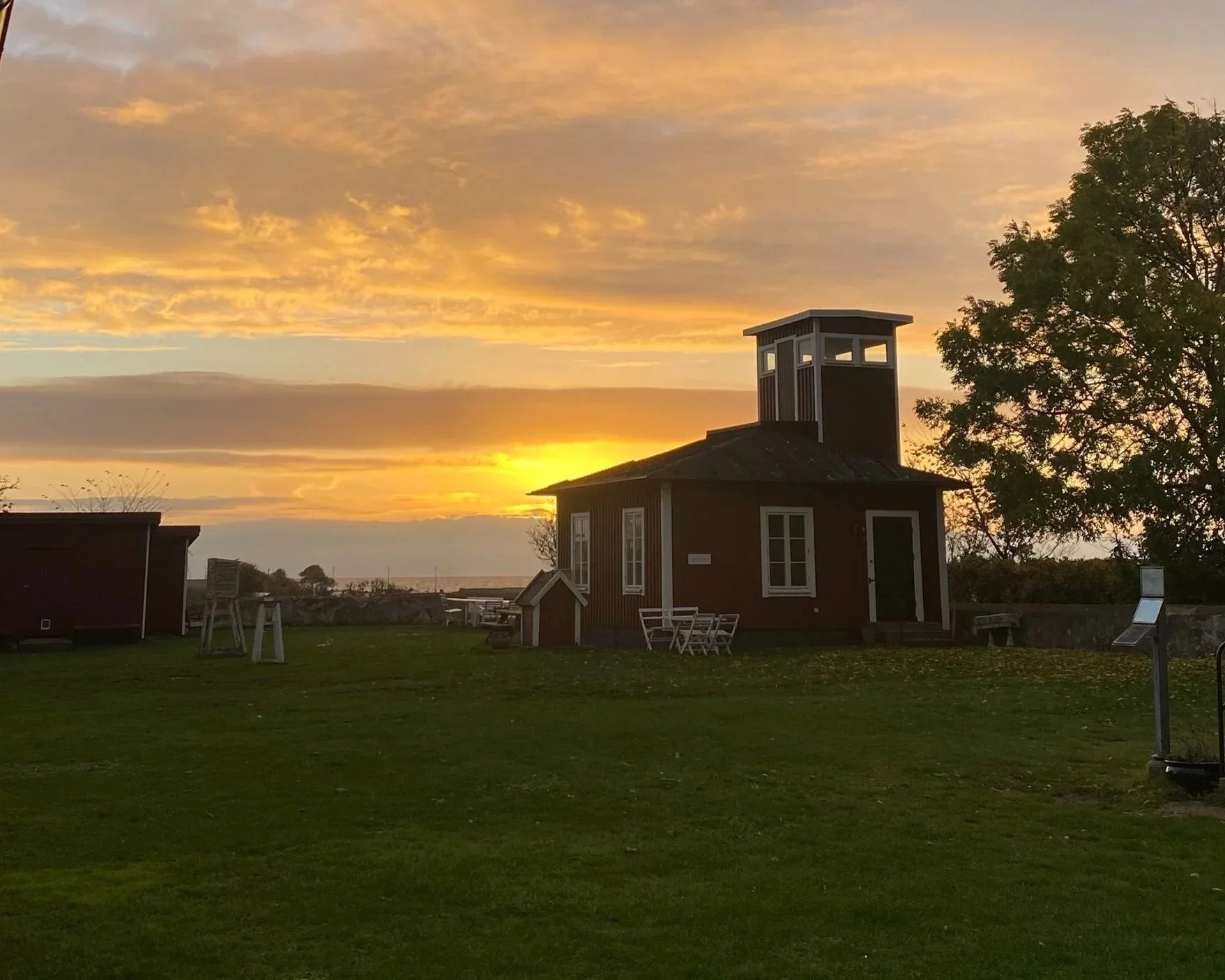 A small red wooden building with white trim and a tower on top, set against a sunset sky with orange and yellow clouds, and a large tree to the right.
