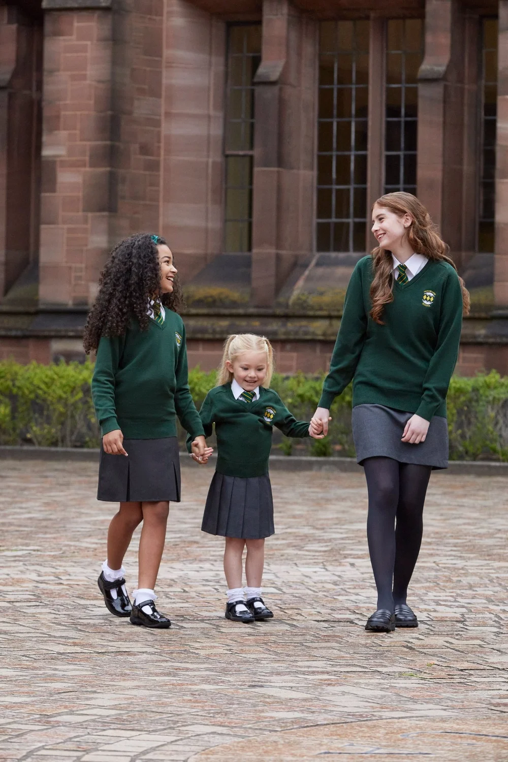 Three pupils in green V-neck jumpers, striped ties and grey skirts walking outside a sandstone school building