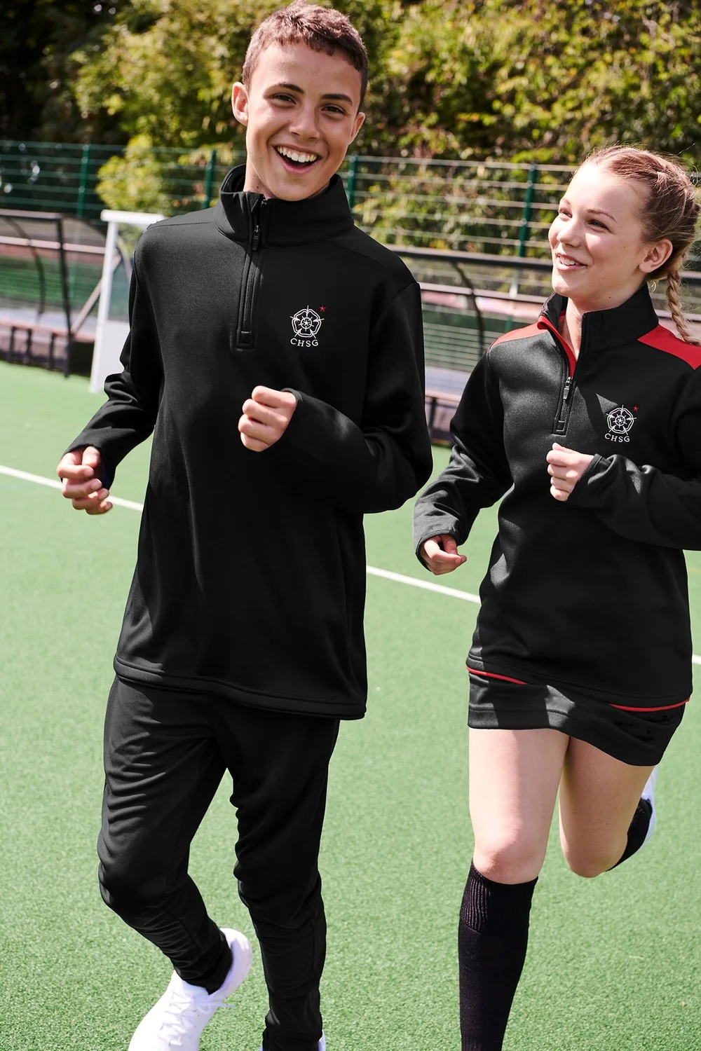 Two pupils in black AKOA quarter-zip midlayers with embroidered school crests running on an all-weather pitch