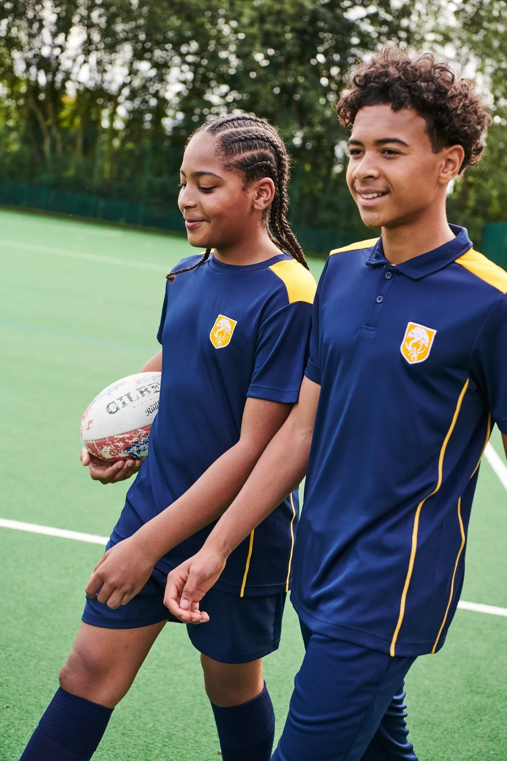 Two pupils in navy and gold PE kit on a sports pitch, one holding a rugby ball