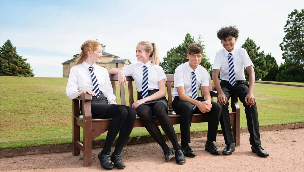 Two pupils in white short-sleeved school shirts, striped navy ties and black trousers, sitting on a bench outdoors in the sunshine
