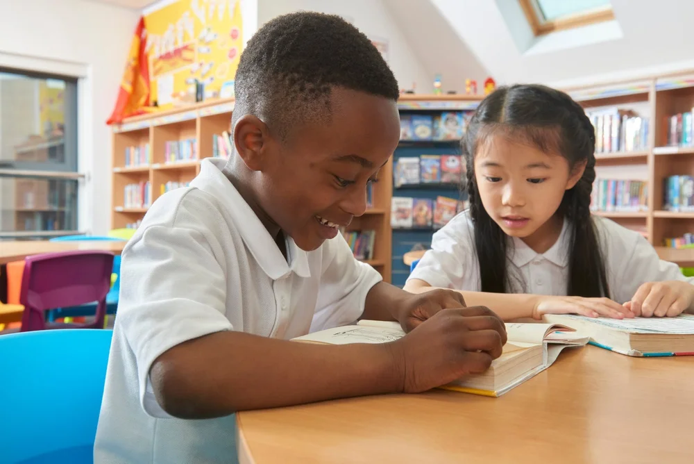 Two pupils in white polo shirts reading books together in a school library