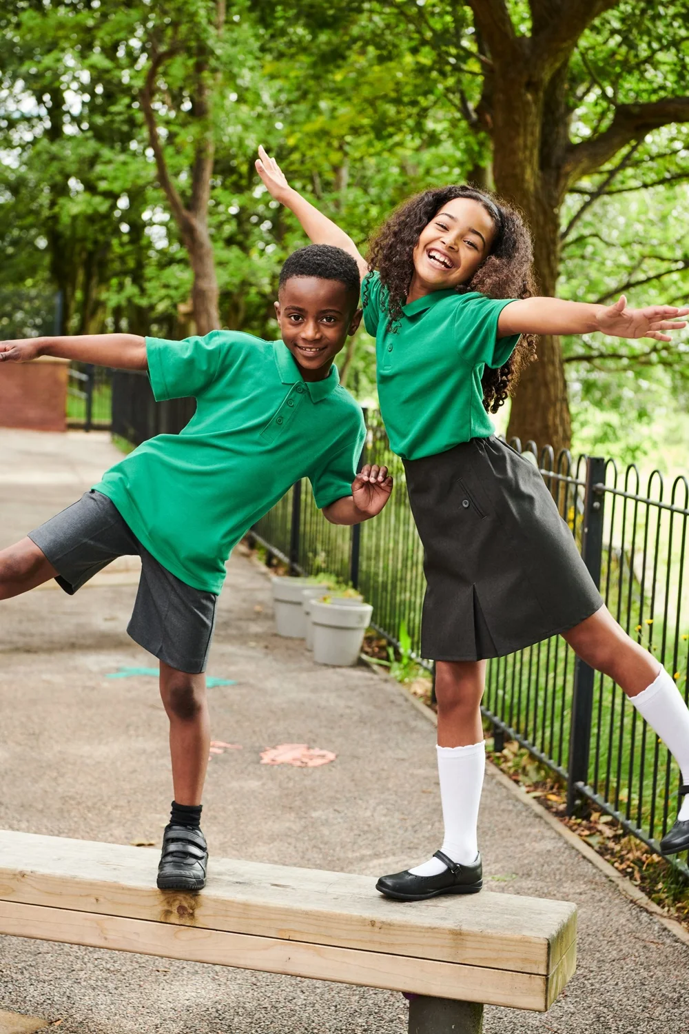 Two pupils in green polo shirts with grey shorts and a grey skirt playing on a log in a park