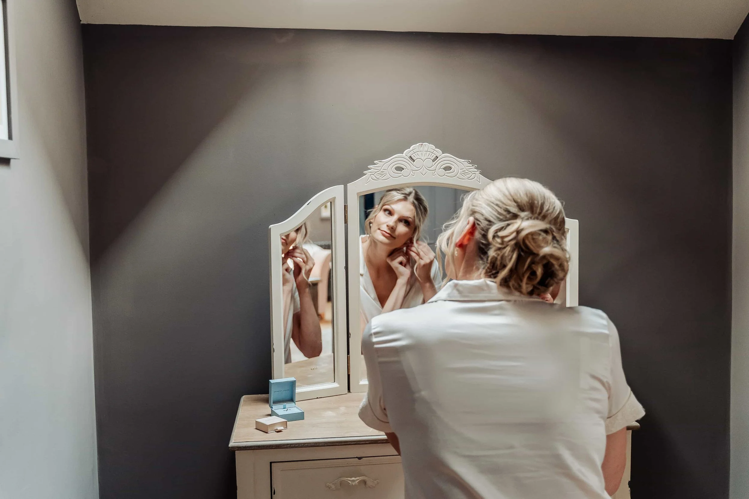 Bride putting in earrings in front of a mirror while getting ready for her Cotswolds wedding
