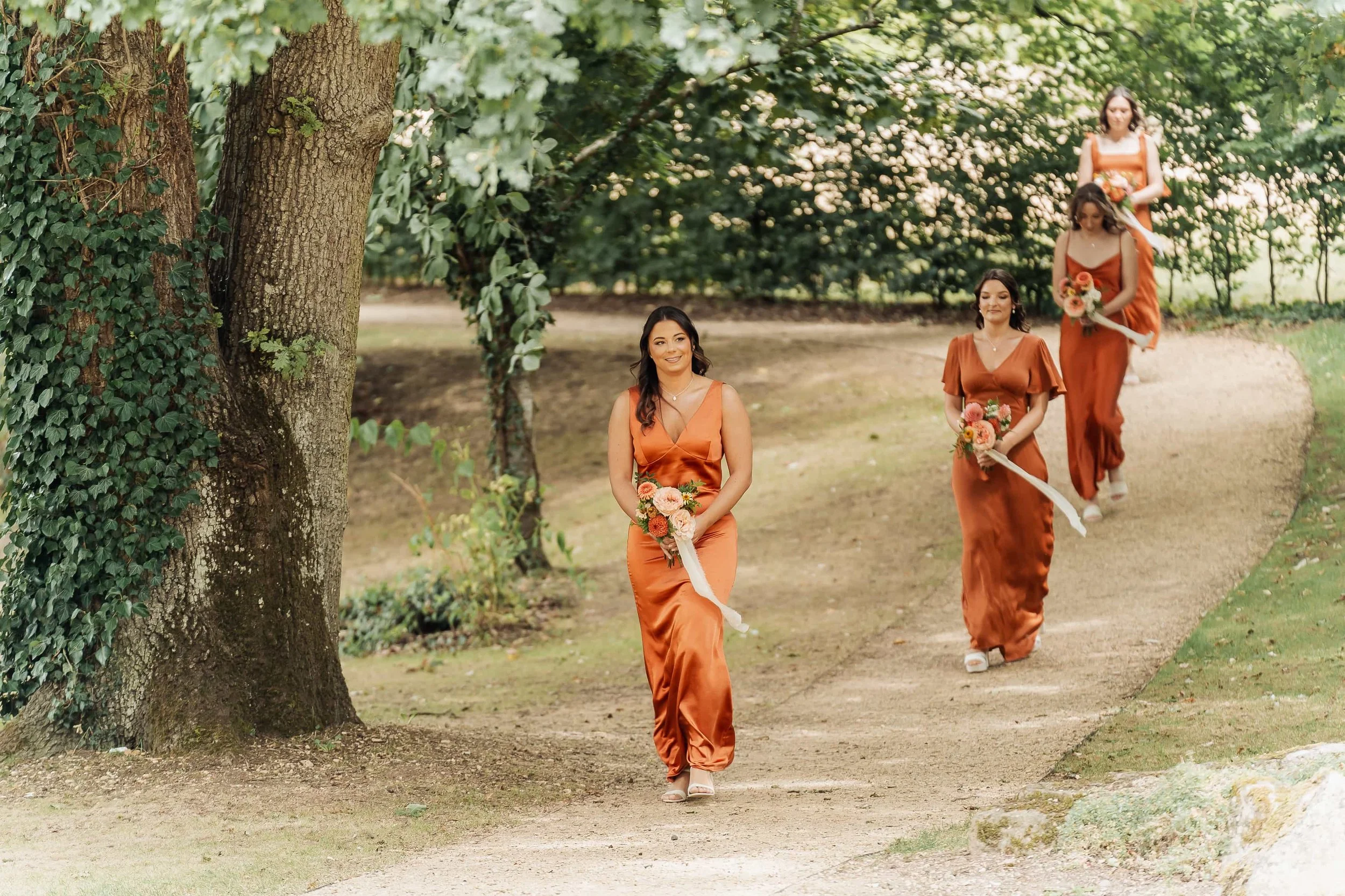 Bridesmaids in burnt orange dresses walking along a tree-lined path at a Cotswolds wedding