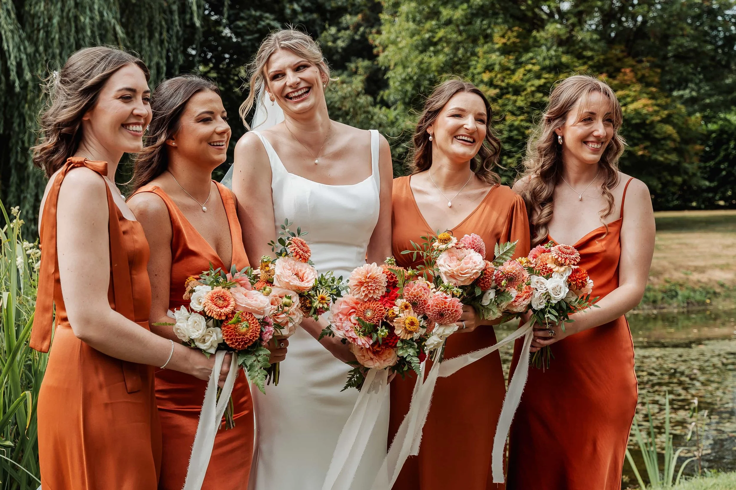 Bridesmaids in orange satin dresses laughing together with willow tree in the background