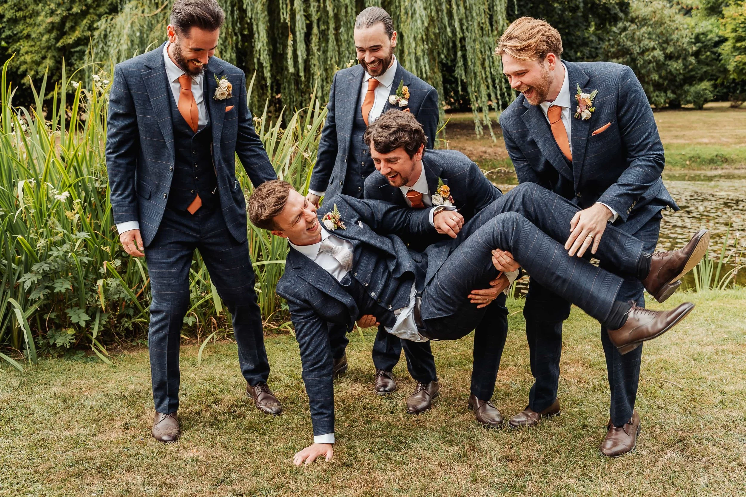 Groomsmen playfully lifting the groom in front of a willow tree by the lake