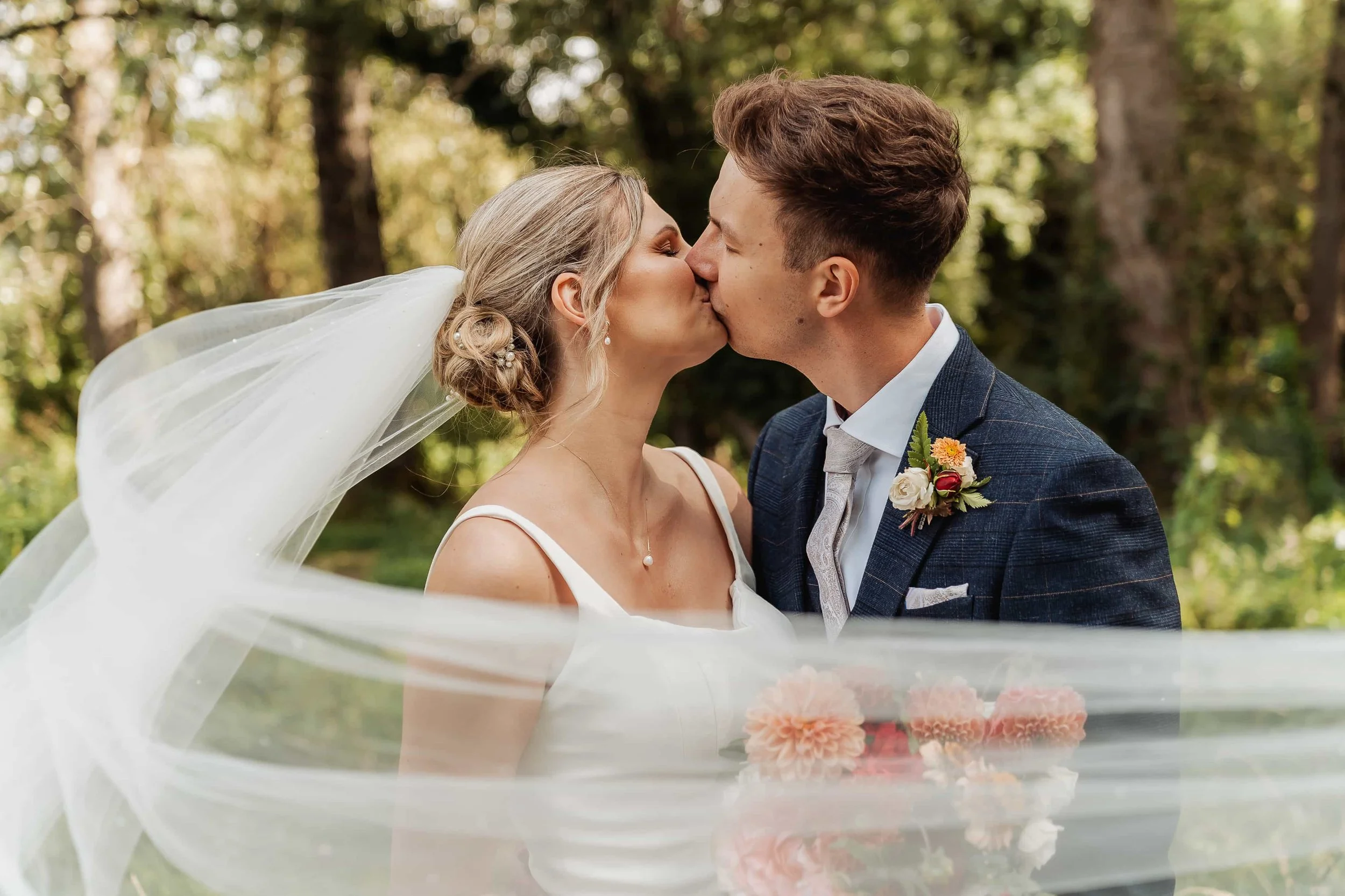 Bride and groom embracing quietly beneath a willow tree with the wedding veil blowing in the wind