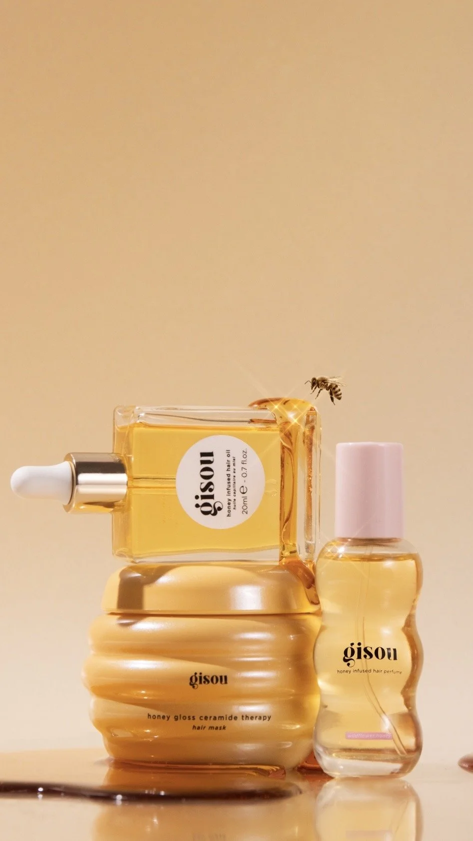 Stacked hair care products including a honey gloss ceramide therapy hair mask, a honey infused hair perfume, and a honey-infused serum with a honeybee flying nearby.