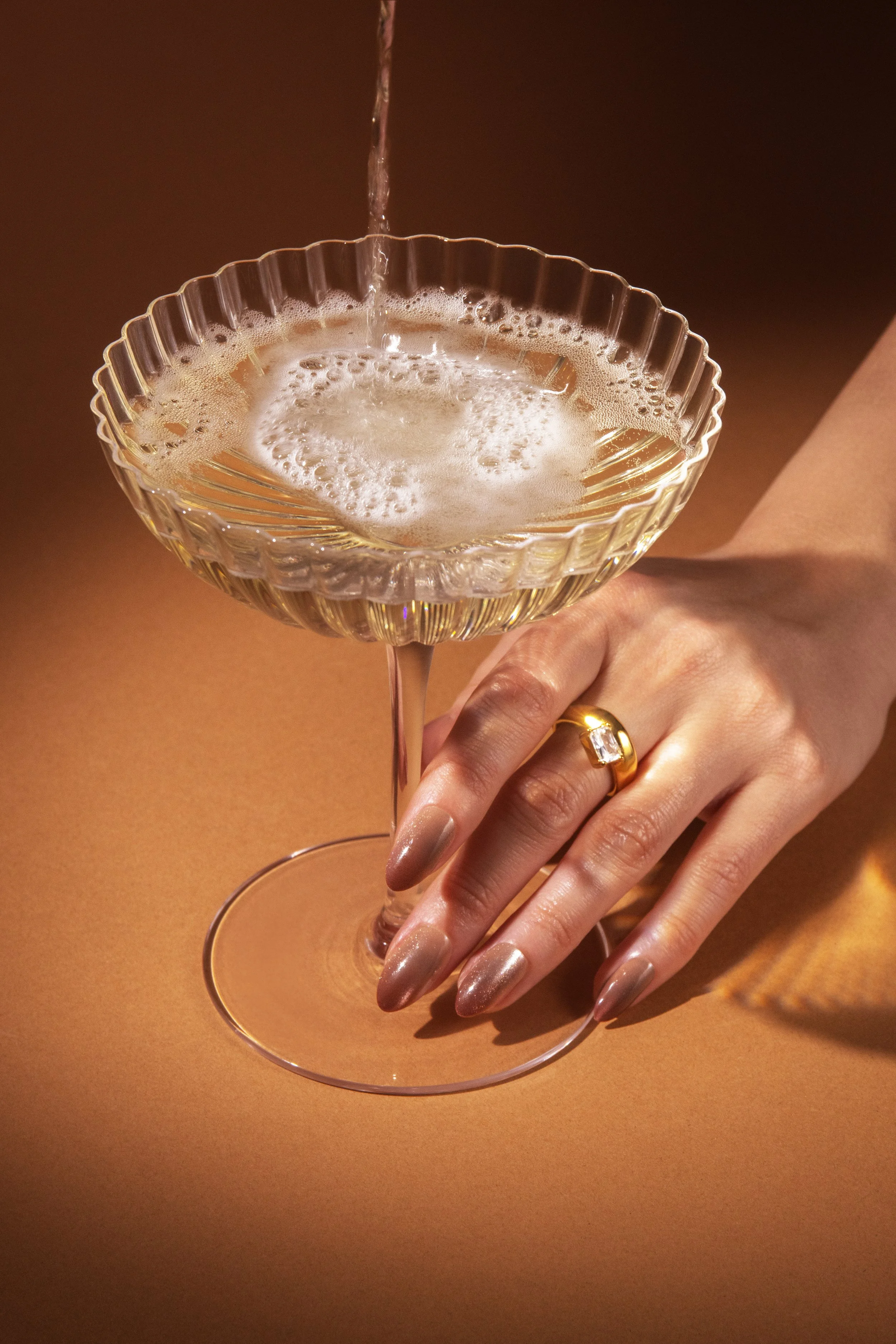 A female hand with a gold ring holding a champagne glass with a decorative pattern, under a stream of bubbling champagne being poured into it.