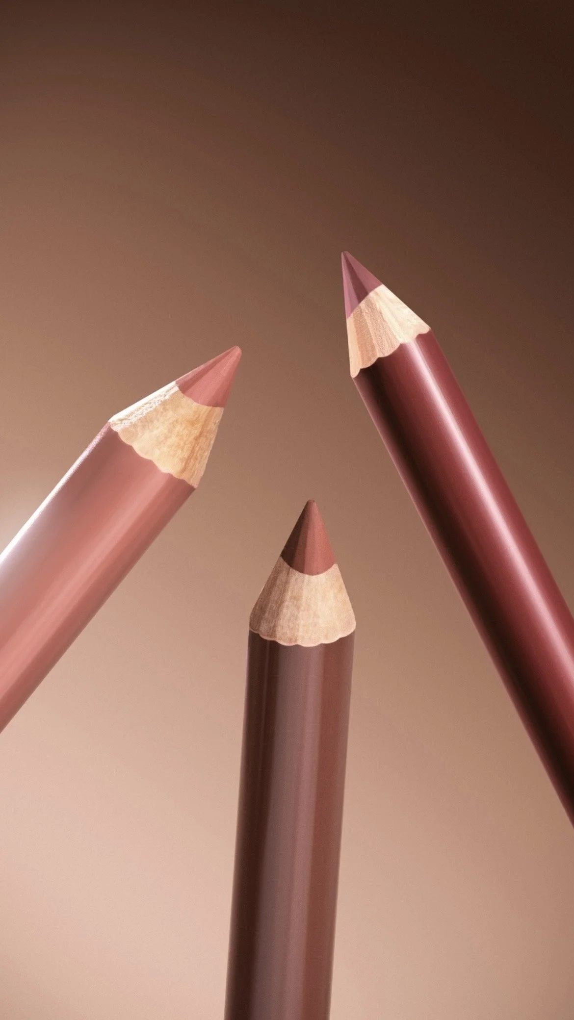 Close-up of three makeup pencils in shades of pink and brown against a beige gradient background.