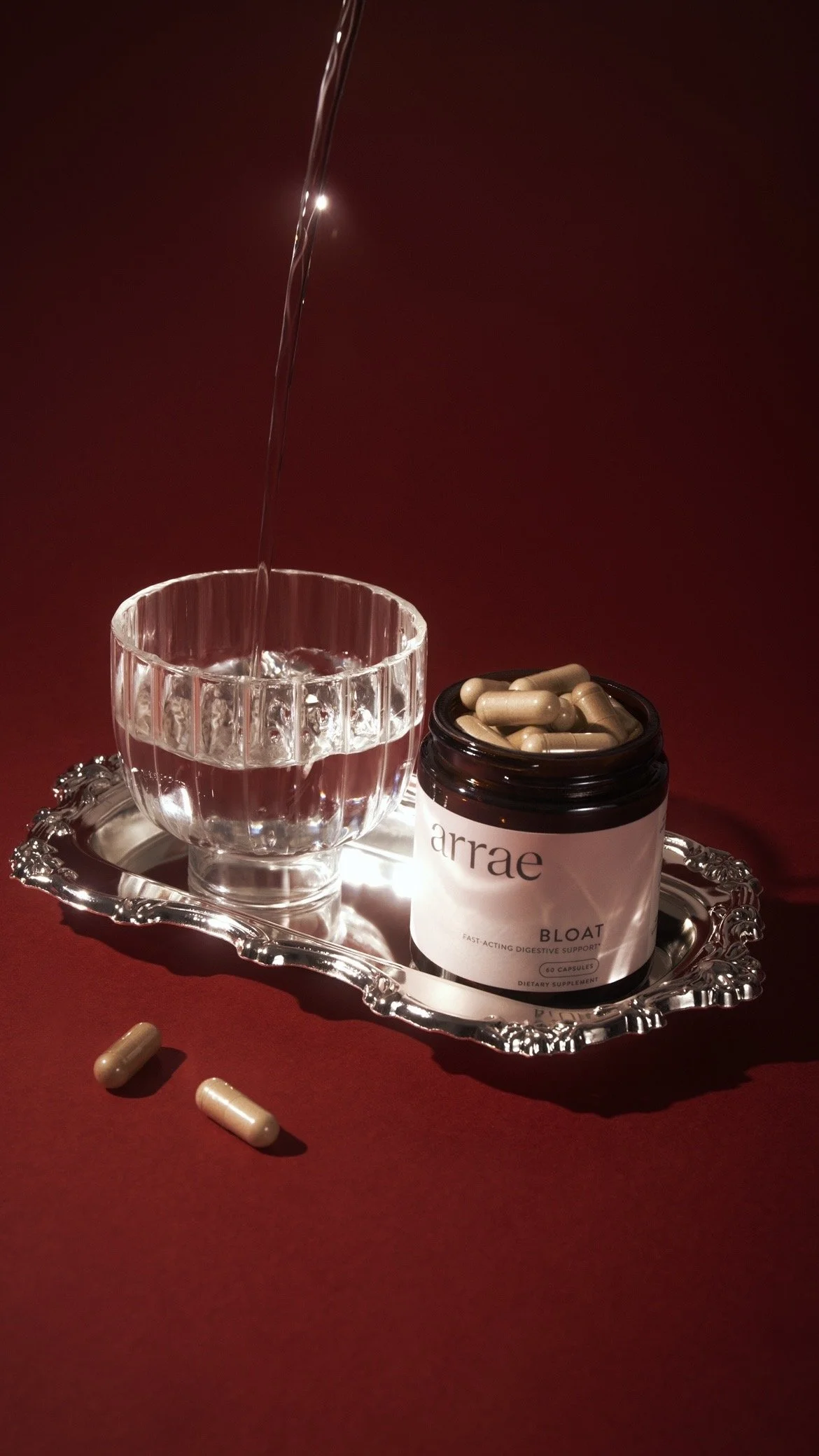A silver tray holding a glass of water, a bottle of dietary supplement capsules labeled 'arrae Bounce,' and a few capsules outside the bottle on a red background.