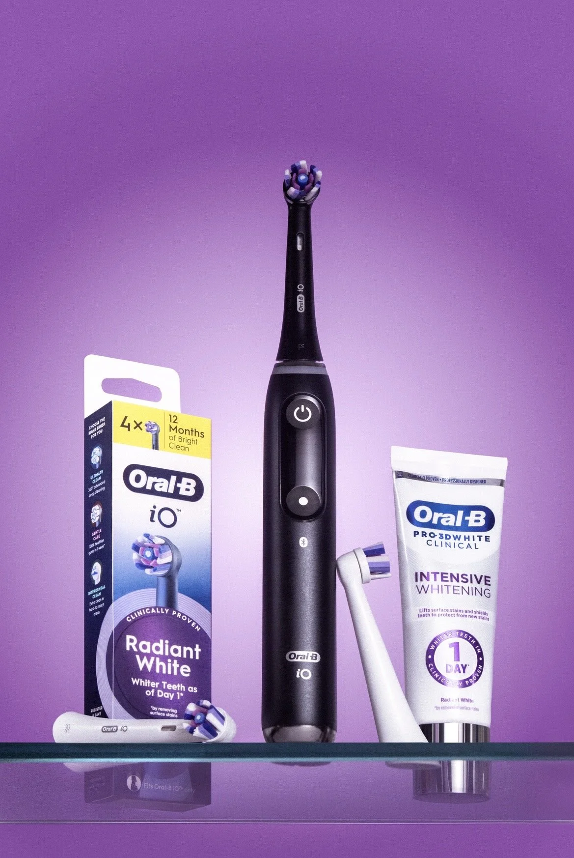 An electric toothbrush with a black handle is positioned upright on a glass shelf, surrounded by a box of Oral-B Radiant White toothpaste, a white toothbrush head, and a tube of Oral-B Pro-3D White clinical intensive whitening toothpaste, all against