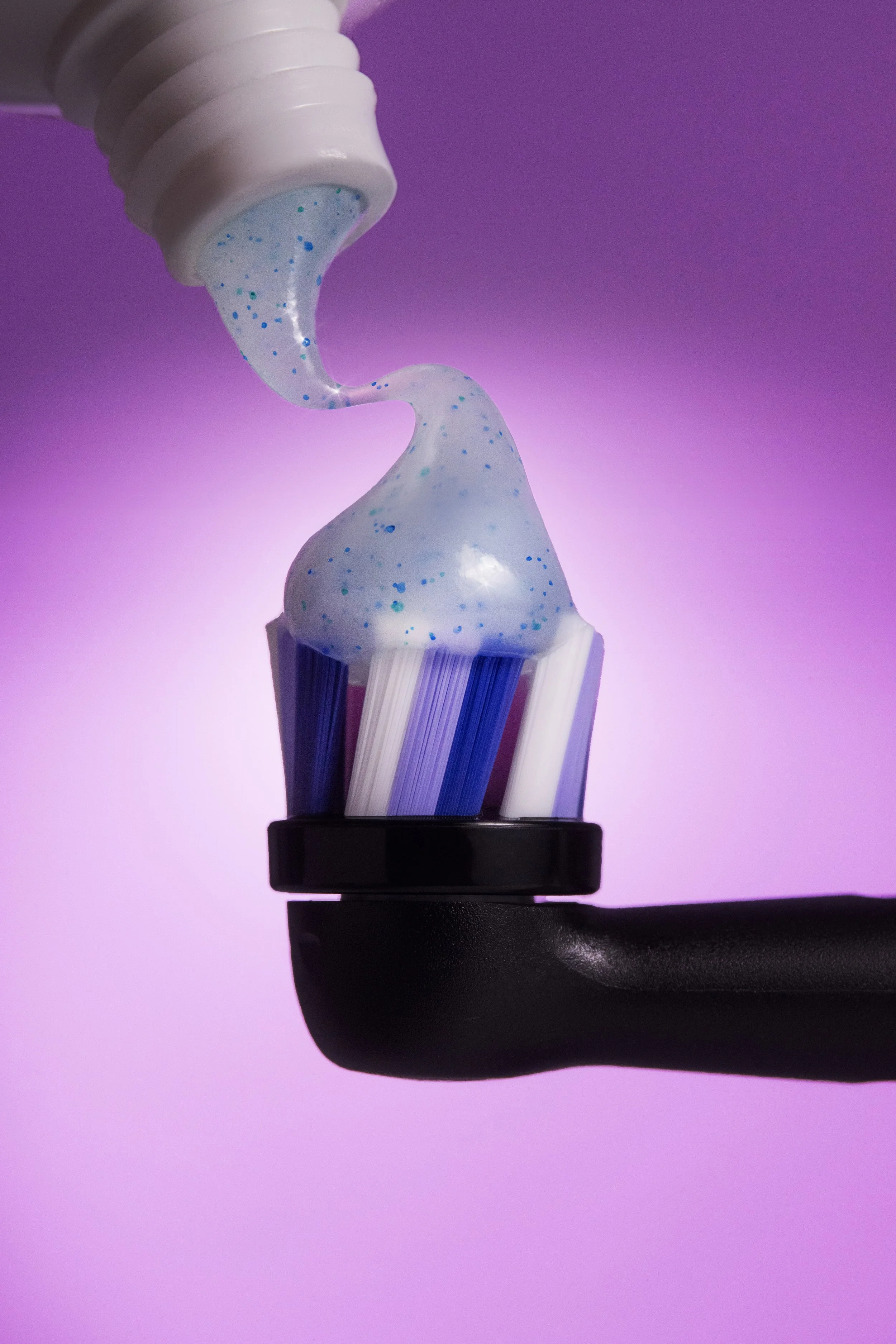 Toothbrush in black handle with blue, white, and purple bristles, with white gel toothpaste dotted with blue on the bristles, against a gradient purple-pink background.