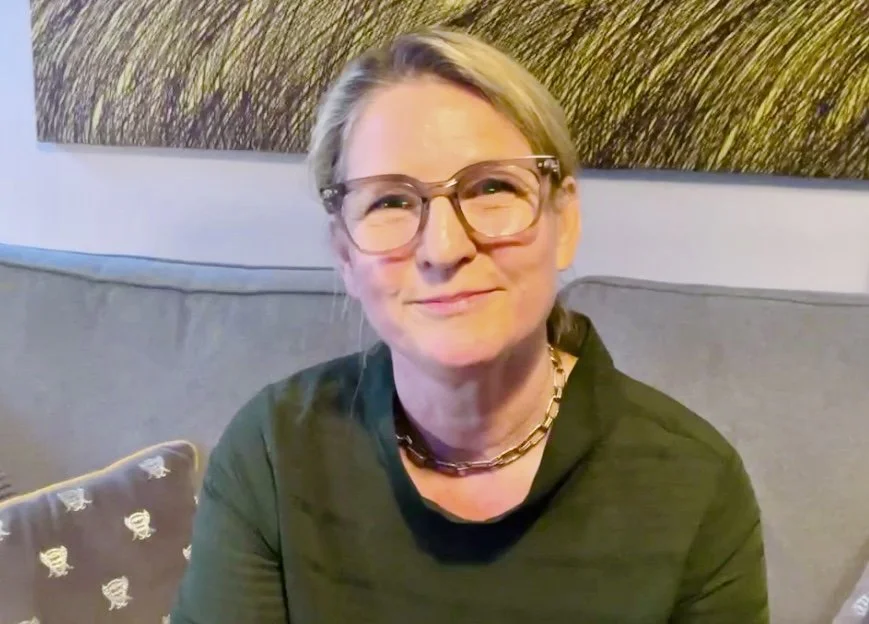 A woman with blonde hair, glasses, and a gold chain necklace sitting on a gray couch, smiling at the camera, with an abstract art piece on the wall behind her.