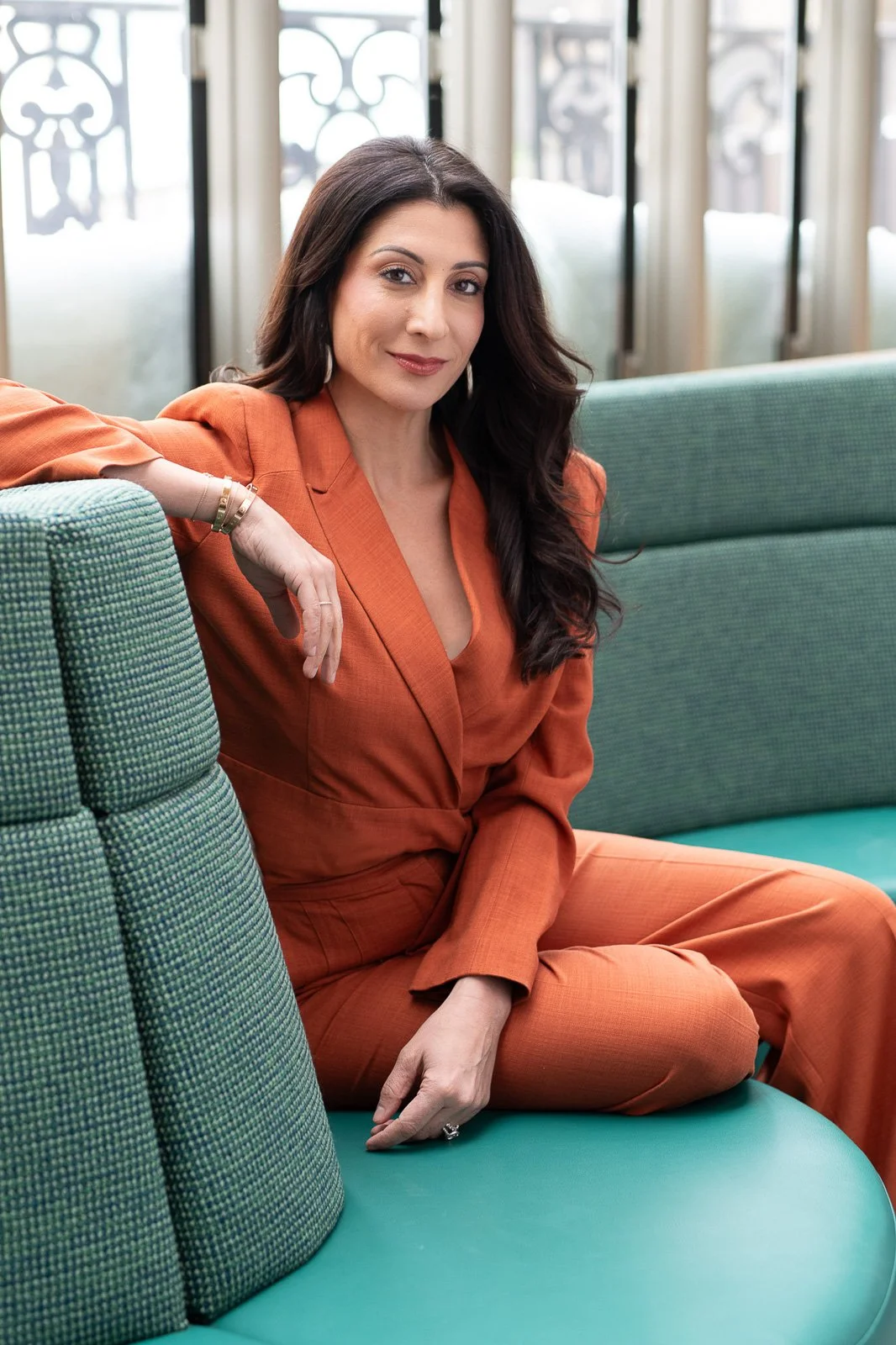 Executive woman with long dark hair and orange jump suit sits on a curved green couch 