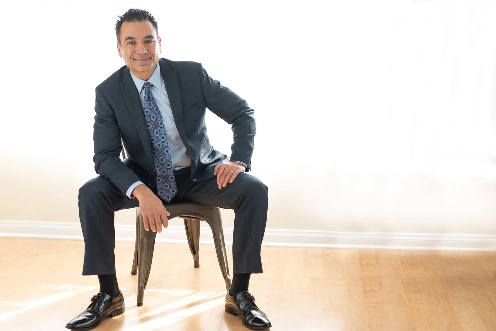 Dark-haired man in blue business suit sitting on chair in room with bright window sheers behind him, during his studio business photoshoot.