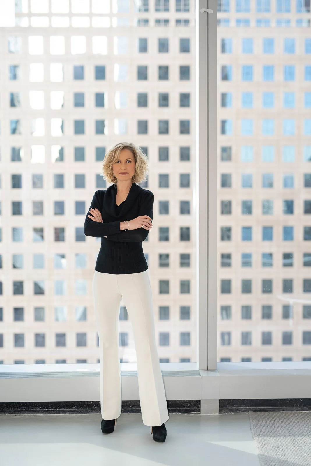 Woman executive in power pose with arms crossed in front of large window with skyscraper windows outside.