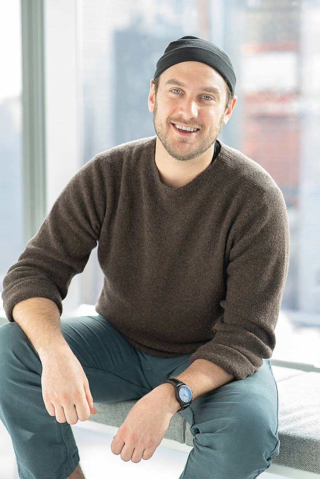 Television chef, Brad Leone wearing a sweater and beanie sits on window seat with cityscape behind him.