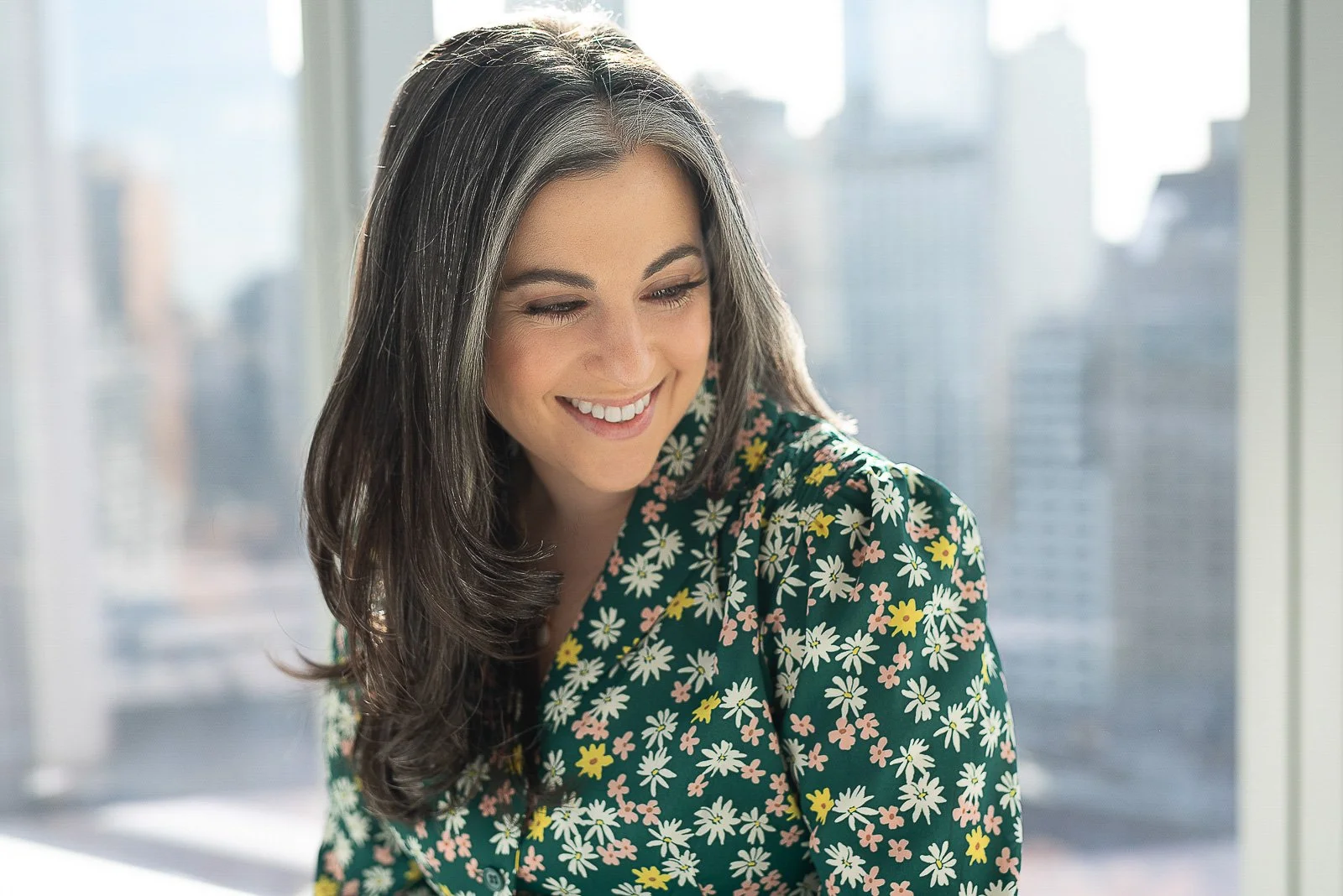 Television chef, Claire Saffitz, wearing a green floral top smiling, looks down to the side with New York City behind her for a promotional photo shoot.