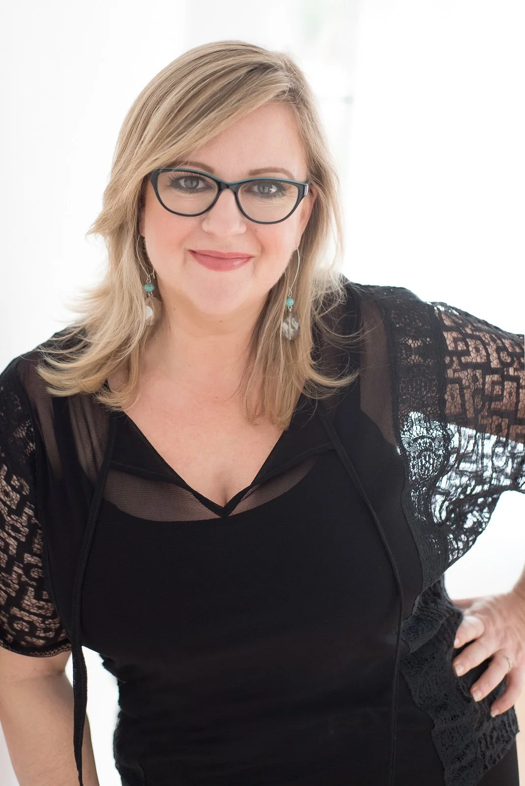 A woman with blonde hair and glasses wearing a black lace dress, standing indoors with a bright, white background.