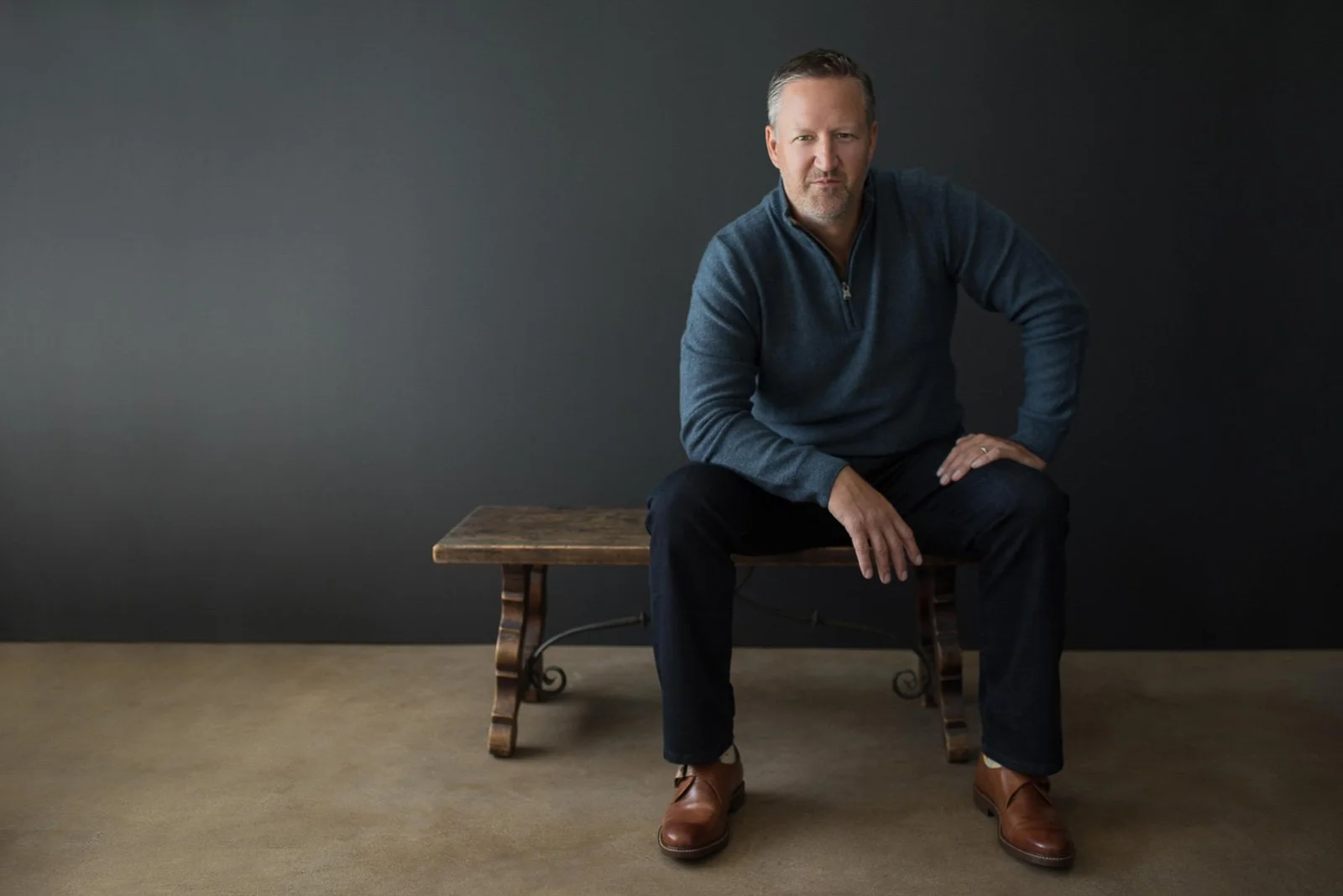 Male entrepreneur seated on wooden bench in dramatic studio lighting for magazine-style portrait.